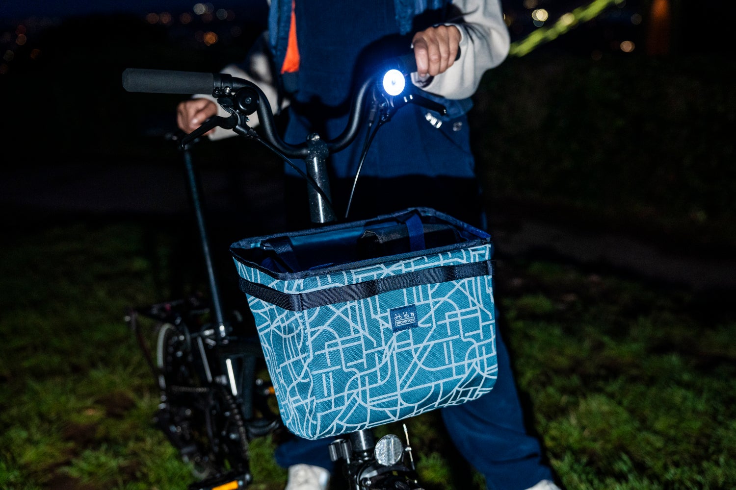 A nighttime shot of the Brompton Bright Nights luggage collection attached to the front of a Brompton folding bike