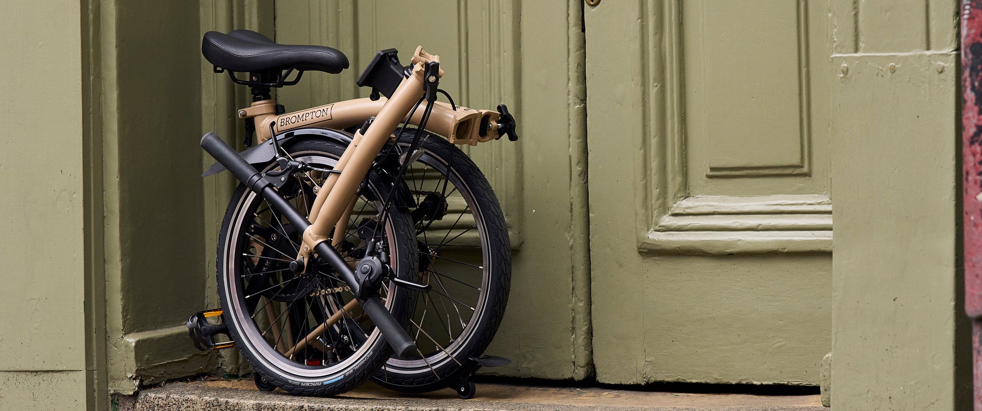 A folded Dune Sands Brompton C Line bike against an earthy neutral door