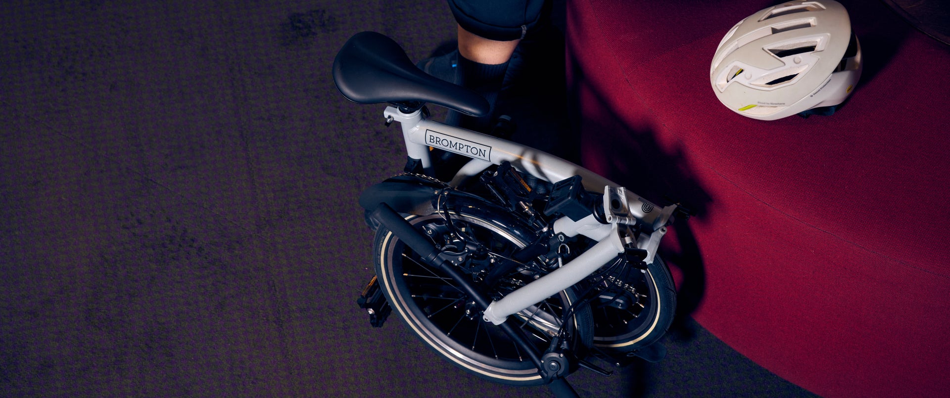 A picture of a folded Lunar Ice Brompton P Line against a burgundy stool and white helmet