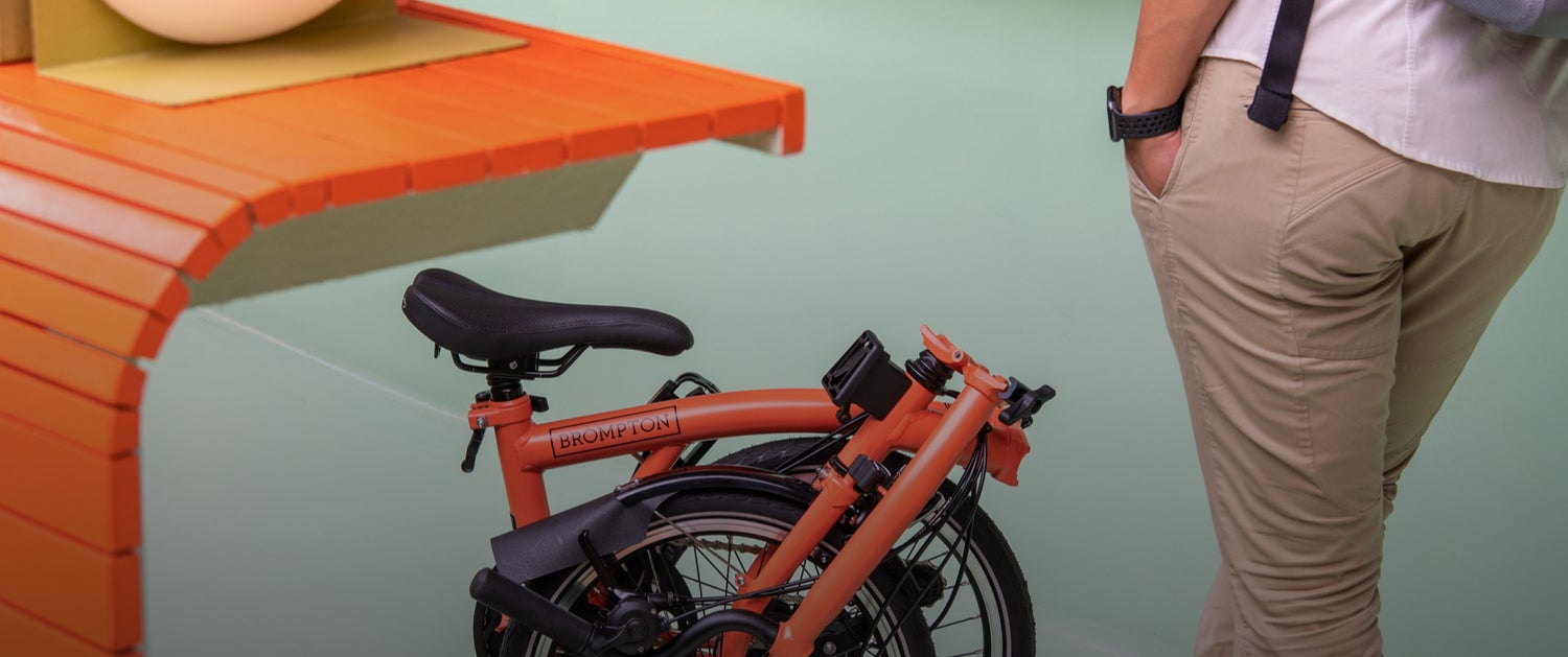 A folded Fire Coral Brompton bike next to an orange table and against a mint green wall