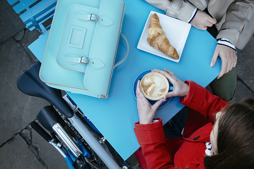 Two people eating crossiants at a table with folded Brompton by their side and Brompton x Cambridge Satchel Company in view