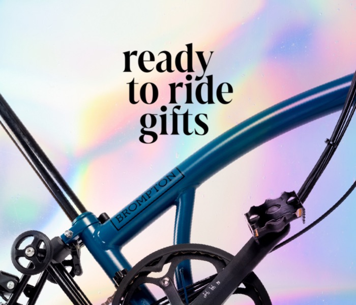 A fun, holographic background with a Brompton C Line in Ocean Blue and Brompton Borough Waterproof Bag Small in the foreground, with graphic text "Ready to ride gifts"